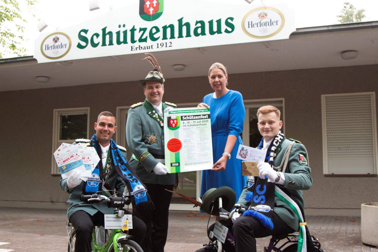 Platzmajor Mark Böhner (von links), Oberst Guido Düsing, Iris Brockmeier (Hauptsponsor Volksbank Schlangen/Spadaka Bad Lippspringe) und Hofoffizier Johannes Klare präsentieren vor dem Schützenhaus die Preise für die Kinder-Tombola.